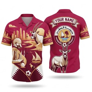 May include: A maroon button-up shirt with a design featuring a skyline, desert landscape, and an oryx. The back of the shirt has a Qatar emblem and space for a name. The shirt has short sleeves and a collar.