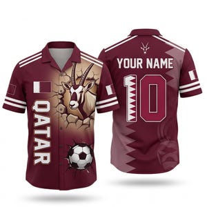 May include: Maroon button-up shirt with white stripes and a Qatar theme. Features a graphic of an oryx breaking through a wall, a soccer ball, and the word "QATAR". The back has the number 10 and space for a name.