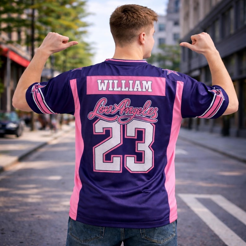 Op de afbeelding: Een marineblauw shirt met roze accenten en de naam "WILLIAM" over de schouders. Het shirt heeft "Los Angeles" in cursief en het nummer "23" in wit. Witte strepen sieren de mouwen.