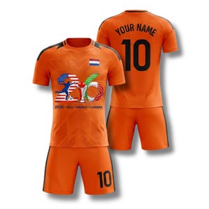 Op de afbeelding: Oranje voetbaltenue met zwarte accenten. Het shirt heeft een afbeelding met de tekst "2026 - USA - MEXICO - CANADA" en het nummer "26". De achterkant van het shirt heeft het nummer "10" en ruimte voor een naam. De shorts hebben ook het nummer "10".