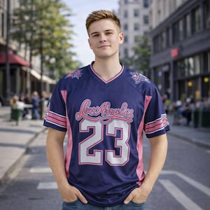 Op de afbeelding: Een marineblauw shirt met roze en witte accenten. Het shirt heeft de tekst "Los Angeles" in een sierlijk lettertype en het nummer "23" in een groot, vetgedrukt lettertype. Het heeft een V-hals en korte mouwen.