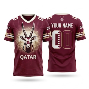 Op de afbeelding: Bordeauxrood shirt met een witte V-hals en gouden details. De voorkant toont een illustratie van een oryx en het woord "QATAR". De achterkant toont "YOUR NAME" boven het nummer "00" met een wit en bordeauxrood ontwerp.