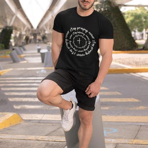 Puede incluir: Camiseta negra con un diseño circular blanco con una cruz y las palabras "Never stop praying, never stop believing". La camiseta se combina con pantalones cortos negros y zapatillas blancas. La imagen está tomada al aire libre.