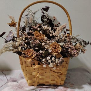 Puede incluir: Una cesta de mimbre llena de flores secas. El arreglo incluye rosas color beige, paniculata blanca y varias hierbas y hojas secas. La cesta tiene un asa y un lazo decorativo.