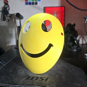 Puede incluir: Una máscara amarilla brillante con forma de cara sonriente, con aberturas para los ojos y la boca. La máscara tiene una superficie lisa y una expresión alegre. Probablemente sea un disfraz o un artículo decorativo.