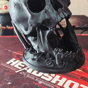 May include: A black, 3D-printed skull sculpture with a spider on top. The skull has detailed features and is set on a round base. The sculpture is on a surface with the text "HEADSHOT" and "AUTO RECOIL SUPPRESSION".