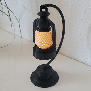 Op de afbeelding: Een zwarte lantaarnlamp met een warme, gele gloed. De lamp wordt ondersteund door een gebogen zwarte standaard en een ronde voet. Links staat een potplant met groene bladeren.