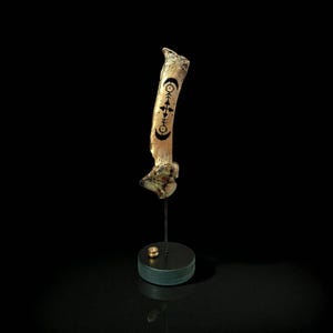 May include: A bone-shaped sculpture with black and gold markings, mounted on a black stand with a gold base. The bone has a natural, off-white colour and features a series of stylized symbols. The background is black, highlighting the sculpture.
