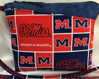 Puede incluir: Un bolso bandolera de tela rojo y azul con cierre de cremallera. El bolso presenta un patrón repetido del logotipo de Ole Miss y las letras "M" en un cuadrado blanco. El bolso tiene una correa negra.