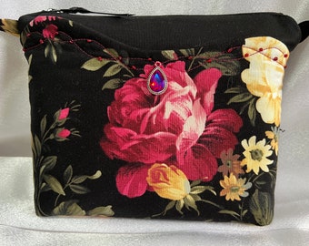 Puede incluir: Una bolsa negra con cremallera con estampado floral que presenta rosas rojas, flores amarillas y hojas verdes. Una joya iridiscente en forma de lágrima está unida a la bolsa.