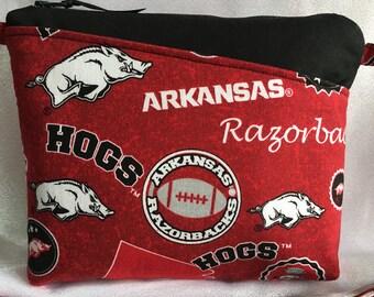 Puede incluir: Bolsa roja con cremallera y forro negro. La tela presenta un patrón repetido de cerdos Razorback blancos, las palabras "Arkansas Razorbacks" y las palabras "Hogs" en negro.