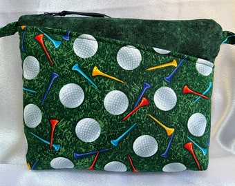 Puede incluir: Una bolsa verde con cremallera con un patrón de bolas de golf y tees. La bolsa tiene un forro de tela verde y un tirador de cremallera negro.