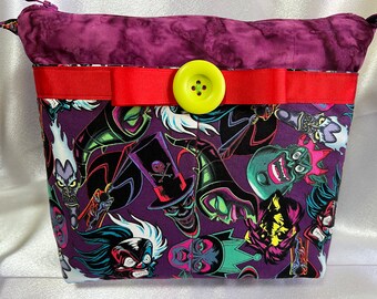 Puede incluir: Bolso de mano de tela morado con una cinta roja y un botón amarillo. El bolso está decorado con una impresión de varios villanos de Disney, incluyendo Ursula, Maléfica y Cruella de Vil.