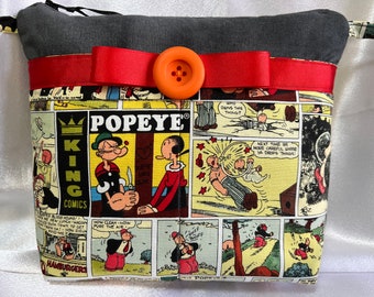 Puede incluir: Una bolsa de tela gris con cremallera, una cinta roja y un botón naranja. La bolsa está decorada con un patrón de cómic colorido con Popeye y el texto "Popeye King Comics".