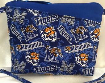 Puede incluir: Un bolso cruzado azul con un estampado del logotipo de los Memphis Tigers, que incluye la mascota del tigre y la palabra "Tigers". El bolso tiene una cremallera y una correa azules, y está hecho de tela.