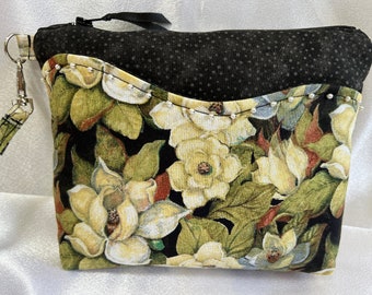 Puede incluir: Una bolsa negra con cremallera con estampado floral que presenta flores blancas de magnolia y hojas verdes. La bolsa tiene un forro de tela de lunares negros y blancos y un tirador de cremallera de cadena plateada.