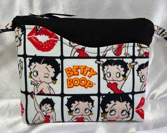 Puede incluir: Bolsa de cremallera negra y blanca con fondo blanco y cremallera negra. La bolsa está decorada con un patrón repetido de imágenes de Betty Boop y las palabras "Betty Boop".