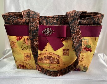 Puede incluir: Una bolsa de mano marrón y burdeos con temática vinícola. La bolsa presenta un lazo burdeos con un adorno plateado y un estampado de uvas, botellas de vino y texto francés. El texto de la bolsa dice "Chateau d'Jeanne" y "Beau Boncoeur".