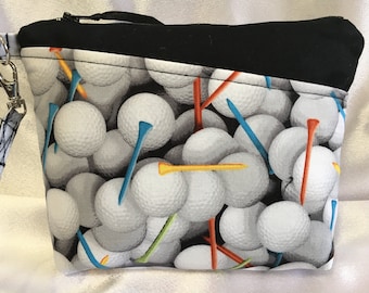 Puede incluir: Una bolsa negra con cremallera con un forro de tela blanco y gris con un patrón de bolas de golf y tees. La bolsa tiene una cremallera negra y una correa negra con un clip plateado.