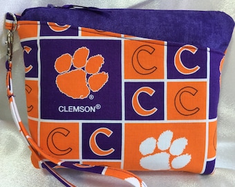 Puede incluir: Una bolsa rectangular con cremallera y correa para la muñeca. La bolsa presenta un patrón repetido de huellas de patas naranjas y la letra "C" sobre un fondo morado. La palabra "CLEMSON" está impresa en la parte delantera.