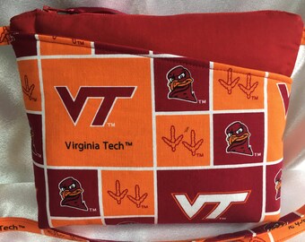 Puede incluir: Un bolso de tela rojo y naranja con cierre de cremallera y una correa para el hombro. El bolso presenta un patrón a cuadros con el logotipo de Virginia Tech, una mascota y las palabras "Virginia Tech".