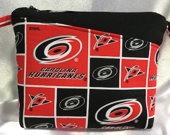 Puede incluir: Una bolsa con cremallera negra y roja con un patrón repetido del logotipo de los Carolina Hurricanes. El logotipo presenta un símbolo de huracán rojo y blanco con las palabras "Carolina Hurricanes" debajo. La bolsa tiene una cremallera negra y una correa negra.