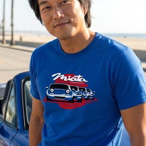 Può includere: Maglietta blu con la scritta "Miata" in bianco e una grafica di auto blu su sfondo rosso. La persona indossa la maglietta vicino a un'auto cabriolet blu, con una spiaggia sullo sfondo.