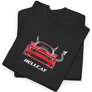 May include: Two black t-shirts are folded and stacked. The top shirt features a red car graphic with the word "HELLCAT" in white. The car has devil horns and a tail. The shirts are likely made of cotton or a similar fabric.