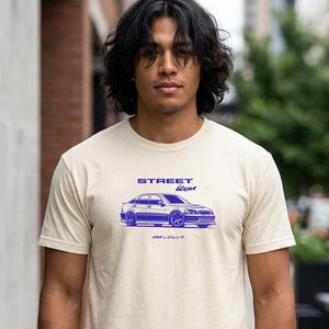 May include: Beige t-shirt with a blue car graphic and the text "STREET ICON". The graphic depicts a sedan with a lowered suspension. The shirt is short-sleeved and made of a soft material.