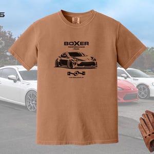 May include: A brown t-shirt with a black graphic of a car and the words "BOXER PERFORMANCE FANS". The shirt also has the text "THE WORLD IS FLAT". The image includes a white, red, and white car in the background, and brown driving gloves.