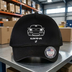 May include: Black trucker hat with a white car graphic and the text "ALLOW POP-UPS". The hat features a mesh back and a curved brim. A circular patch with the American flag and text "PRINTED & FULFILLED IN THE USA" is on the side.