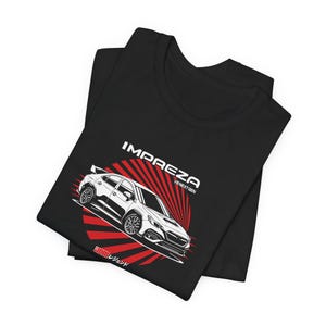 May include: A black t-shirt featuring a white graphic of a car with the text IMPREZA V8 NEXT GEN and JDM followed by Japanese characters. The car is positioned over a red sunburst design. The shirt is folded to display the front graphic clearly.