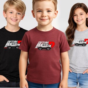 May include: Three children wearing t-shirts with a car graphic. The shirts are black, burgundy, and gray. Each shirt features the text "INITIAL D" with a white car illustration and a red "D" design.