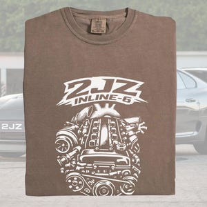 May include: Brown t-shirt featuring a white graphic of a car engine and the text "2JZ INLINE-6". The shirt is folded and has a tag at the top. A car is visible in the background.