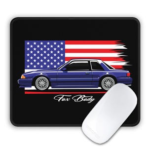 Könnte beinhalten: Schwarzes Mauspad mit einer blauen Autografik mit der amerikanischen Flagge im Hintergrund. Der Text "Fox Body" befindet sich unter dem Auto. Eine weiße Computermaus befindet sich im Vordergrund.