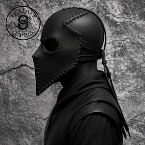 May include: A black leather plague doctor mask with stitched detailing and eye holes. The mask features a long, beak-like nose and is secured with straps. The Leather Supply logo is visible. The mask is worn by a person.