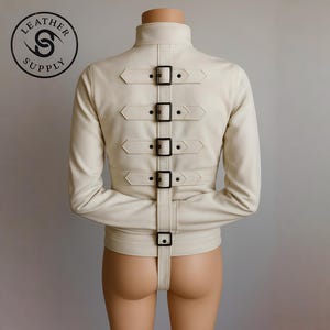 May include: Cream-colored leather jacket with a high collar and long sleeves. The back features five horizontal straps with black buckles and a vertical strap. The Leather Supply logo is visible in the upper left corner.