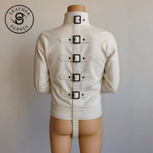 May include: Cream-colored leather jacket with a high collar and multiple black buckle straps down the back. The jacket has long sleeves and a strap that extends from the lower back. The Leather Supply logo is visible in the upper left corner.