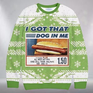 Może przedstawiać: Zielona i biała świąteczna bluza z napisem "I GOT THAT DOG IN ME" nad grafiką hot doga. Bluza ma zielony kołnierz i mankiety oraz powtarzający się wzór płatków śniegu i drzew.