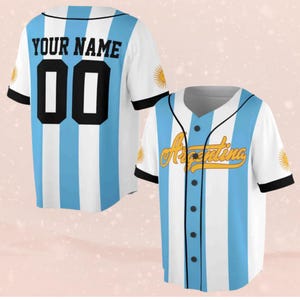 Könnte beinhalten: Zwei Baseball-Trikots, weiß mit hellblauen vertikalen Streifen und schwarzen Verzierungen. Eines hat "YOUR NAME" und die Nummer "00" auf dem Rücken. Das andere hat "Argentina" in goldener Schrift auf der Vorderseite.