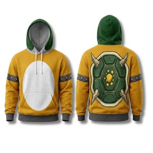 Op de afbeelding: Een gele hoodie met een groene capuchon, een grote witte ovaal op de voorzijde en een donkergrijze zoom. De mouwen hebben donkergrijze banden met zilveren studs. De achterkant toont een grote afbeelding van een groen schildpadschild.