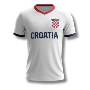 May include: White Croatia football shirt with a red and blue trim on the collar and sleeves. The word "CROATIA" is printed in blue across the chest. A Croatian coat of arms is on the upper left chest.