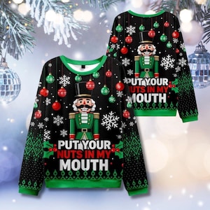 Op de afbeelding: Zwarte kersttrui met een groen en wit ontwerp. De trui heeft een notenkraker-afbeelding en de tekst "PUT YOUR NUTS IN MY MOUTH". Rode en groene ornamenten en sneeuwvlokken zijn ook aanwezig.