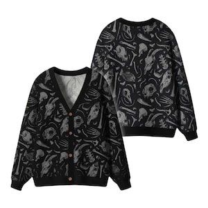 Könnte beinhalten: Schwarze Strickjacke mit V-Ausschnitt und Knopfverschluss, mit einem sich wiederholenden Muster aus grauen Schädeln und Knochen. Ein passender Rundhals-Pullover ist im Hintergrund zu sehen, der ebenfalls das gleiche Knochen- und Schädeldesign zeigt. Die Strickjacke hat eine lockere Passform.