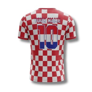 May include: A red and white chequered football shirt featuring the number 10 and the text "YOUR NAME" in white. The number 10 has a blue and red colour scheme. The shirt has short sleeves.