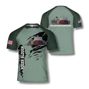 Może przedstawiać: Jasnozielona koszulka z grafiką frachtowca z Wielkich Jezior. Koszulka ma ciemnozielone rękawy i amerykańską flagę na rękawie. Tekst "ROGER BLOUGH" jest wydrukowany pionowo z przodu.