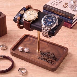 May include: A wooden watch display stand with two watches, a bracelet, rings, earrings, and a pen holder. The stand has the name "Matthew" engraved on it. A stack of books and a decorative glass sphere are also present.