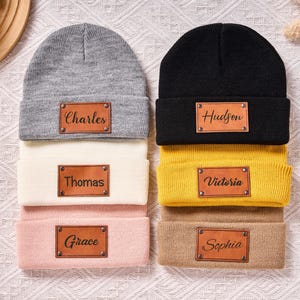 May include: A stack of six beanies in various colors: gray, cream, pink, black, yellow, and tan. Each beanie has a rectangular leather patch with a name: Charles, Thomas, Grace, Hudson, Victoria, and Sophia. The beanies are on a white patterned surface.
