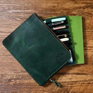 Puede incluir: Un estuche de cuero verde oscuro con cremallera, que contiene varios bolígrafos de varios colores. El estuche está abierto, revelando un cuaderno verde. La palabra "Jackson" es visible en el estuche.