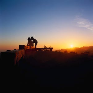 May include: A silhouetted scene of two people on a bench, watching a sunset. The sky is a gradient of blue to orange and yellow. A dark, wooden structure is in the foreground.